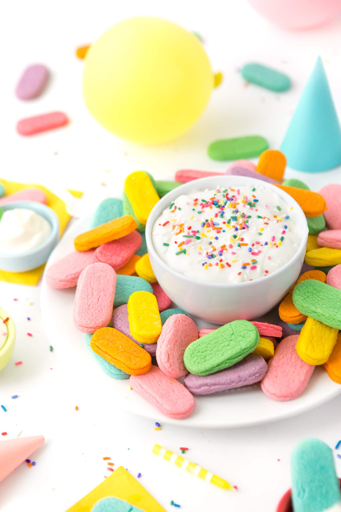 Plate of rainbow sprinkle sugar cookies with a bowl of cream cheese frosting in a party scene.