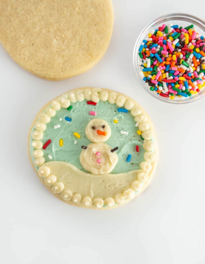 blue snowglobe cookie with white snowman in center, decorated with rainbow sprinkles