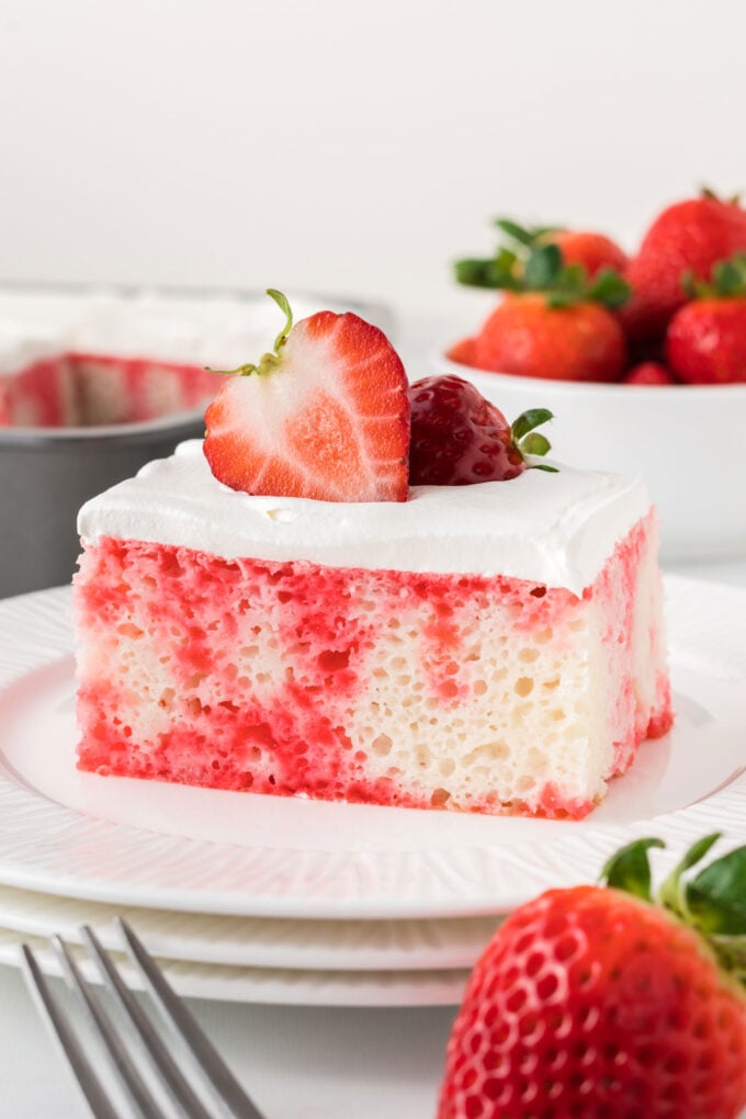 Piece of strawberry Jello poke cake garnished with strawberries on white plate with cake and strawberries in background